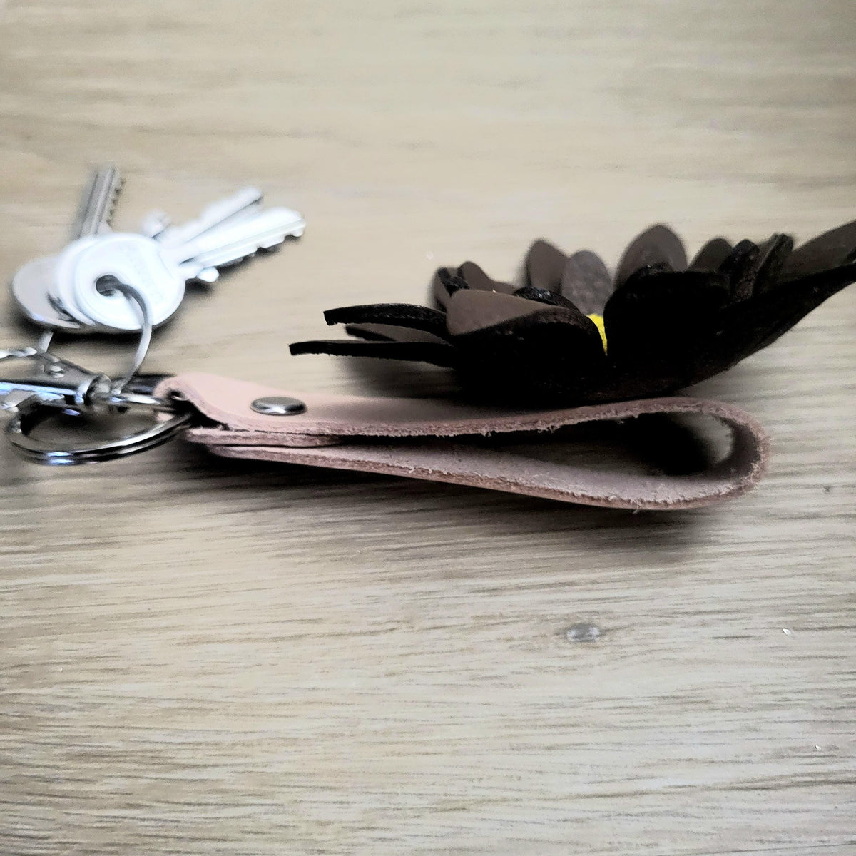 A keychain with a  brown leather loop, a dark brown flower-shaped charm, and silver metal keys on a light wood surface.